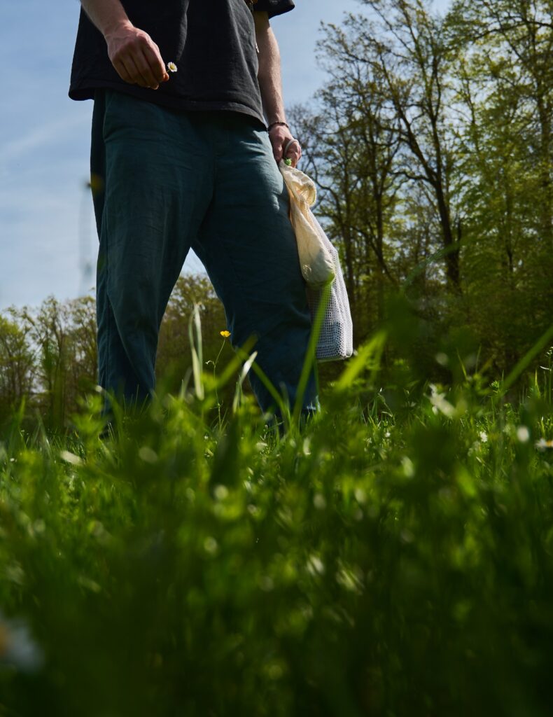 Der Fokus des Bildes liegt auf den Beinen und dem Sammelsäckchen des Mannes der gerade über eine grüne Wiese läuft mit einem Gänseblümchen in der Hand